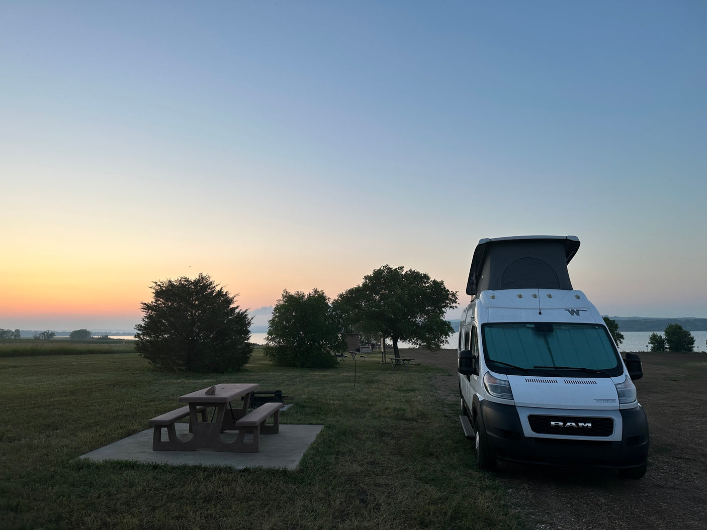 An RV rental camper van parked at a tranquil lakeside campsite with a picnic table, under a beautiful sunrise sky.