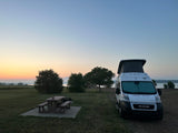 An RV rental camper van parked at a tranquil lakeside campsite with a picnic table, under a beautiful sunrise sky.