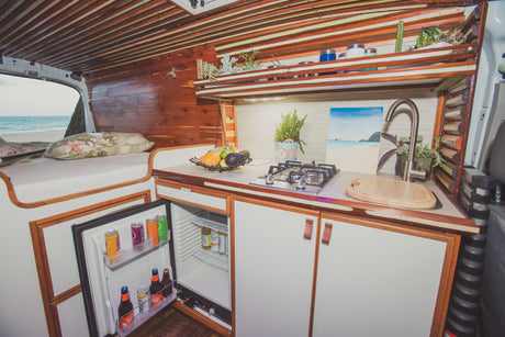 Inside a custom camper van: cozy bed, full kitchen with fridge, and stunning beach view, ideal for an RV rental getaway.