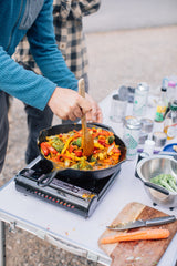 Hands stir a vibrant vegetable stir-fry on a portable stove. Ideal outdoor cooking setup for your RV rental trip!