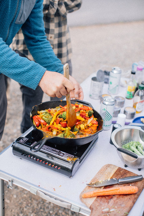 Hands stir a vibrant vegetable stir-fry on a portable stove. Ideal outdoor cooking setup for your RV rental trip!