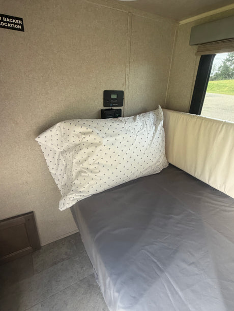 RV rental interior showing a bed with gray sheets, patterned pillow, and wall-mounted controls. A window offers a view of green landscape.