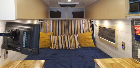 Step inside this comfortable RV rental camper van, featuring a cozy bed, wall-mounted TV, smart storage, and warm wood accents.