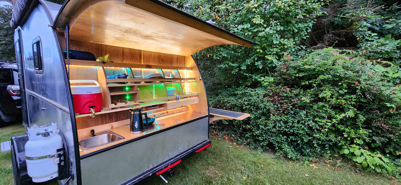 RV rental: Teardrop trailer's illuminated outdoor kitchen with sink, cooler, and propane tank, ideal for camping.