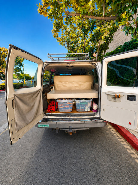 A custom white camper van with its rear doors open, showing a bed, storage bins, and a roof rack. Great RV rental for trips!