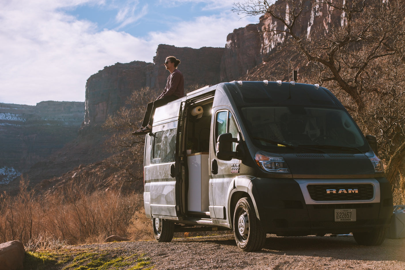 Person sits on dark gray RV rental van roof, admiring vast desert canyons. Open door shows interior. Ideal for van life adventure.