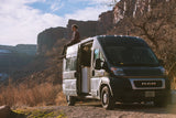 Person sits on dark gray RV rental van roof, admiring vast desert canyons. Open door shows interior. Ideal for van life adventure.