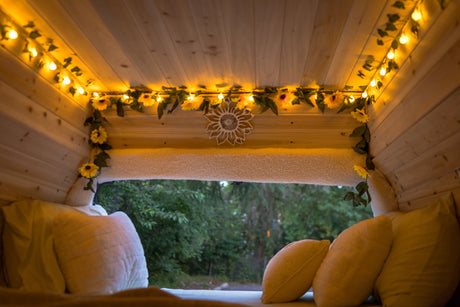 Cozy RV rental interior with warm lights, sunflower garlands, and a nature view. Relax on the comfy bed inside this charming camper.