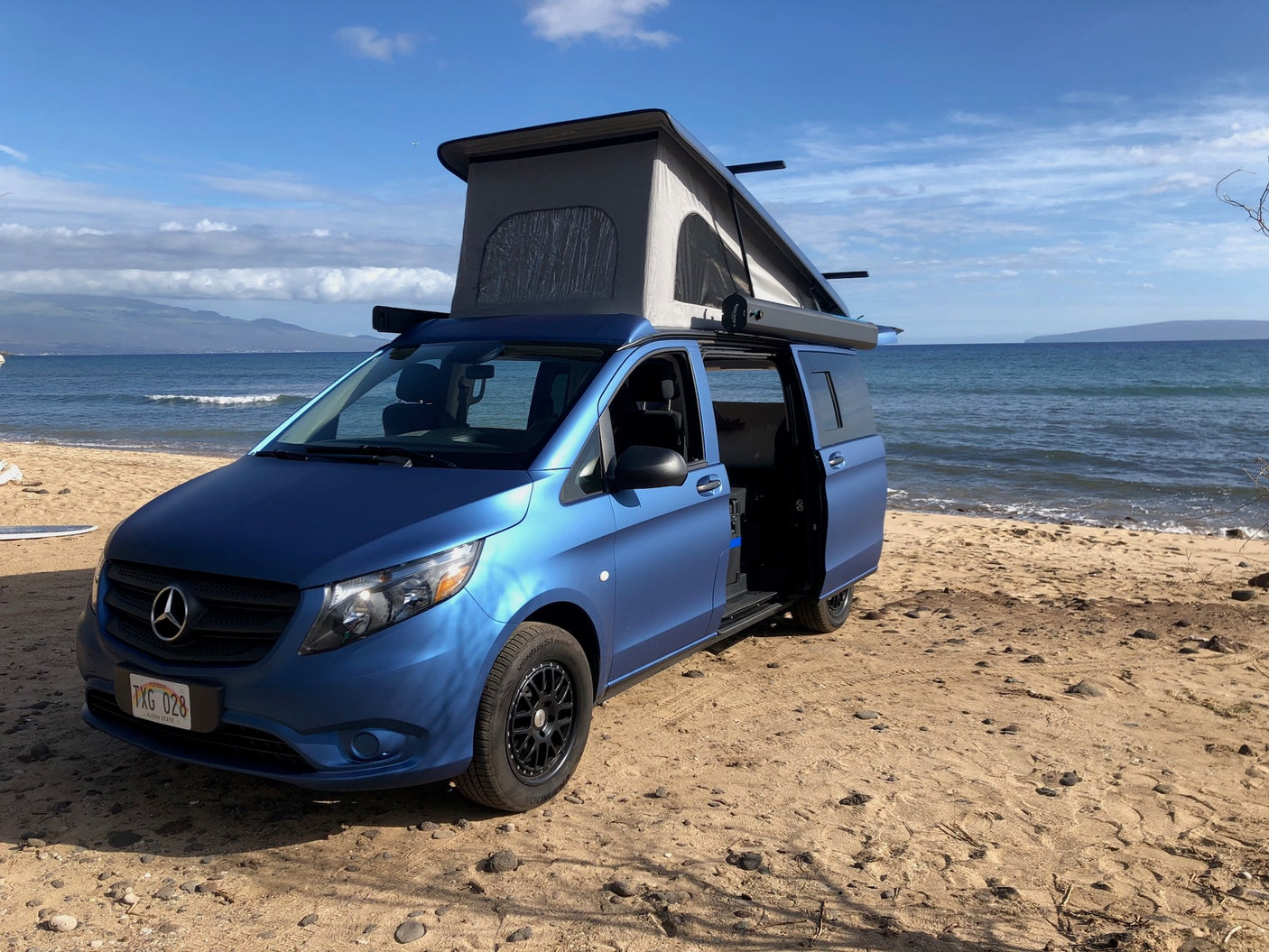 Blue camper van RV rental on a sunny beach with pop-top open & ocean views. Perfect for your next adventure!