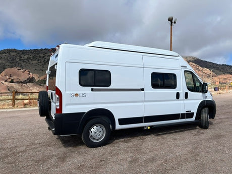 White Winnebago Solis camper van for rv rental parked on gravel, backed by mountains and a cloudy sky. Ideal for adventure travel.