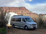 A sleek grey rv rental camper van parked at a desert campsite, with a white dome tent and towering red rock cliffs.