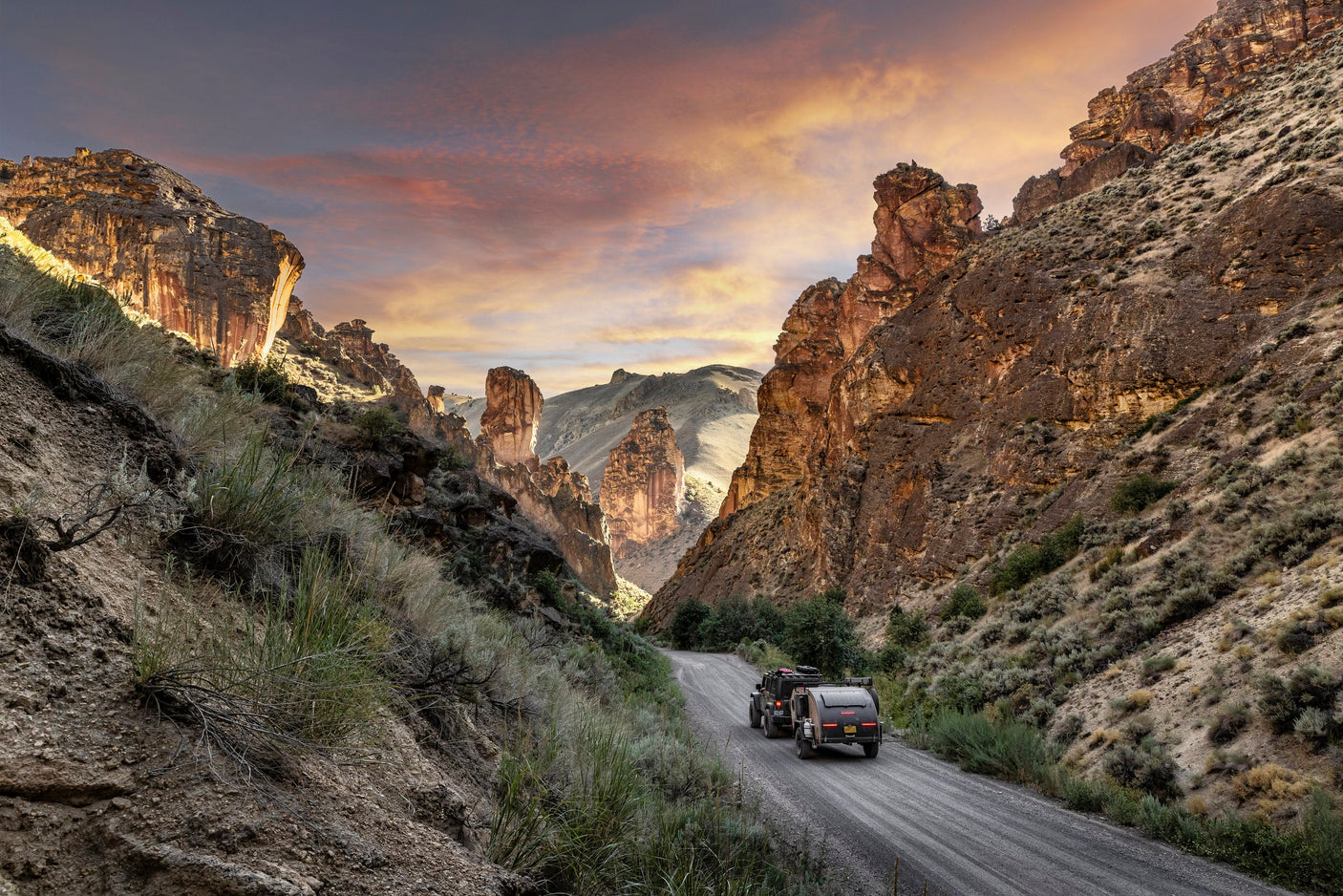 An RV rental rig travels a rugged canyon road at sunset, flanked by majestic rock formations.