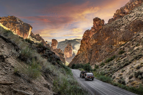 An RV rental rig travels a rugged canyon road at sunset, flanked by majestic rock formations.