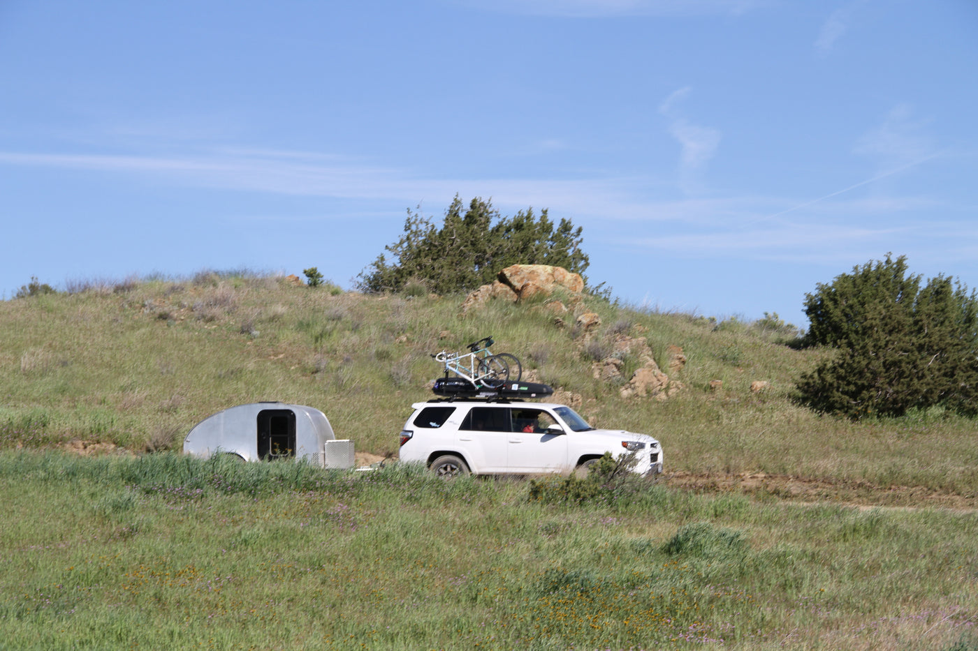 A white SUV with a roof-mounted bike tows a compact silver RV rental trailer on a dirt road through grassy hills. — photo 15