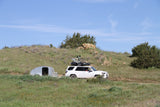 A white SUV with a roof-mounted bike tows a compact silver RV rental trailer on a dirt road through grassy hills.