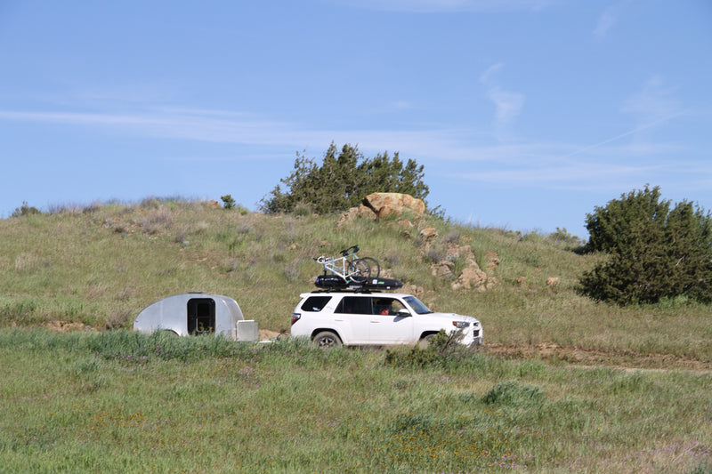 A white SUV with a roof-mounted bike tows a compact silver RV rental trailer on a dirt road through grassy hills. — photo 15