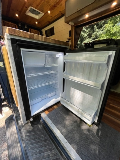  and open fridge.|Inside this RV rental: open compact refrigerator showing freezer and fridge shelves. Perfect camper van kitchen setup.|Explore this bright camper van RV rental interior! Features wood walls — photo 6