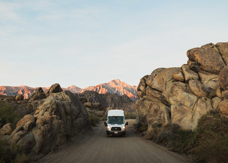 Adventure awaits: White RV rental van drives on a dirt road through dramatic rock formations with mountains at sunset.