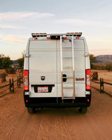 Rear view of a white Ram ProMaster camper van with ladder & roof rack, parked on sandy desert road. Perfect for your next RV rental adventure!