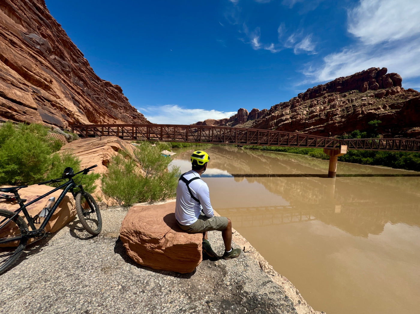 A cyclist rests by a muddy river, with a bike, bridge, and red rock cliffs under a blue sky, ideal for an RV rental journey. — photo 19