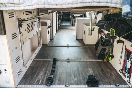 An RV rental's versatile interior shows modular white storage, L-track flooring, and various gear secured under a bed.