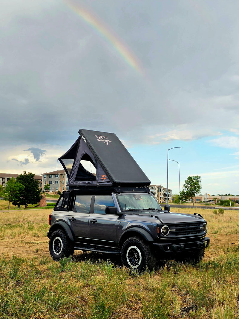  parked by hills and a highway overpass.|Gray Ford Bronco with an open rooftop tent in a grassy field. An ideal RV rental for off-road camping and outdoor adventure.|A Ford Bronco with a rooftop tent — photo 3