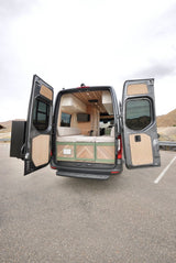 Rear doors of a gray camper van are open, revealing a bright custom interior with wood details and green accents. Perfect RV rental.