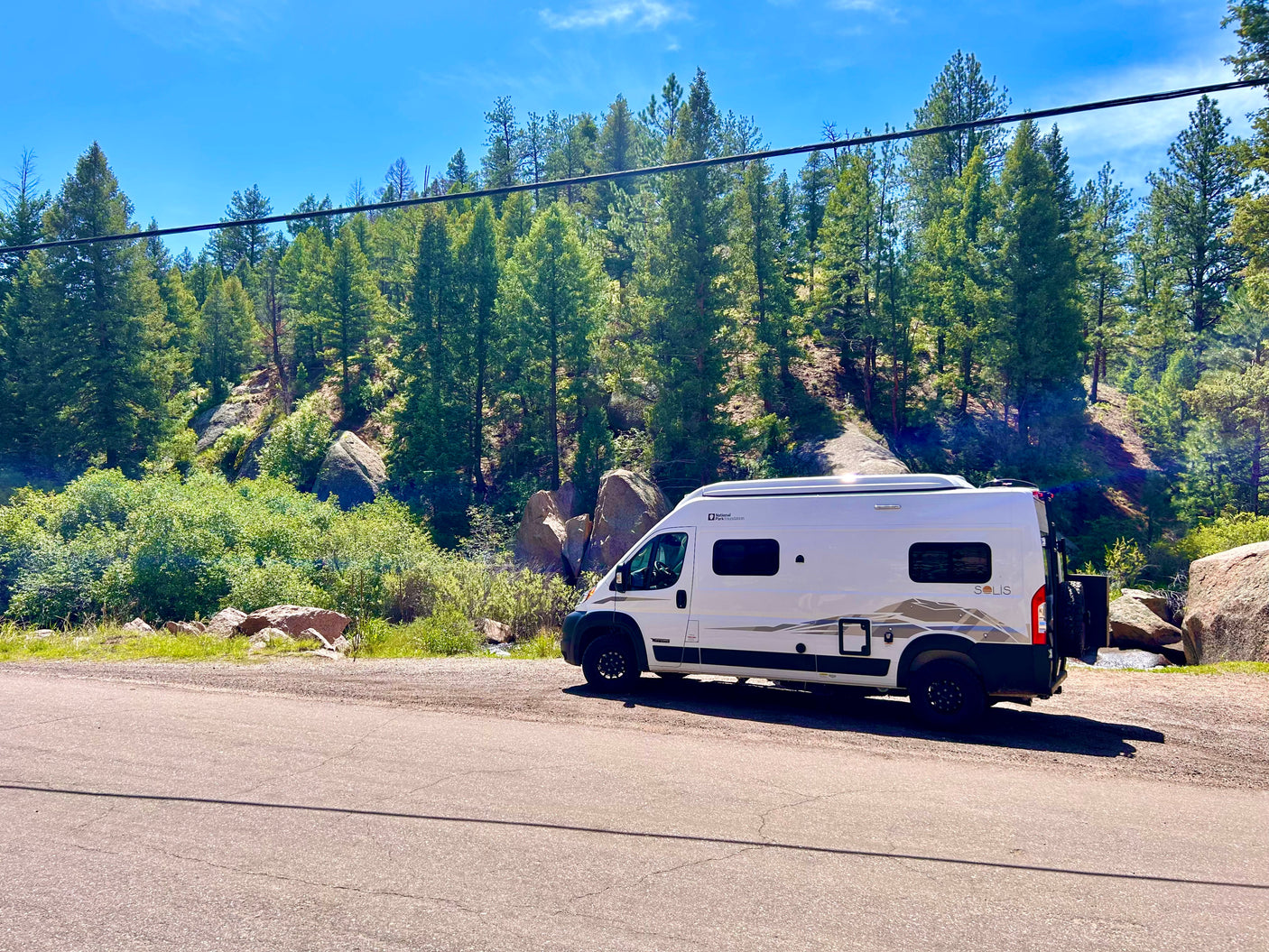 White Winnebago Solis RV rental parked on a mountain road. Sunny forest, rocks, and blue sky create a scenic backdrop.