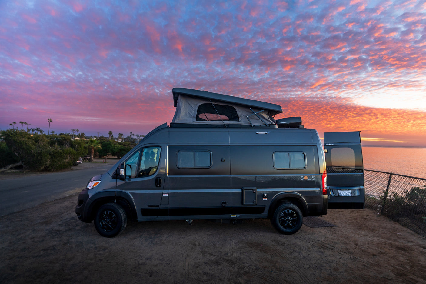 A gray Winnebago Solis camper van (RV rental) with its pop-top extended, parked oceanfront under a vibrant sunset sky.