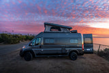 A gray Winnebago Solis camper van (RV rental) with its pop-top extended, parked oceanfront under a vibrant sunset sky.