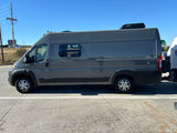 Grey RV rental camper van with a Dometic roof unit, parked on concrete, ready for travel and towing a trailer.