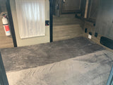 RV rental interior: two grey-blanketed bunks, a fire extinguisher, white curtain, and wood-paneled walls. Cozy sleeping space.