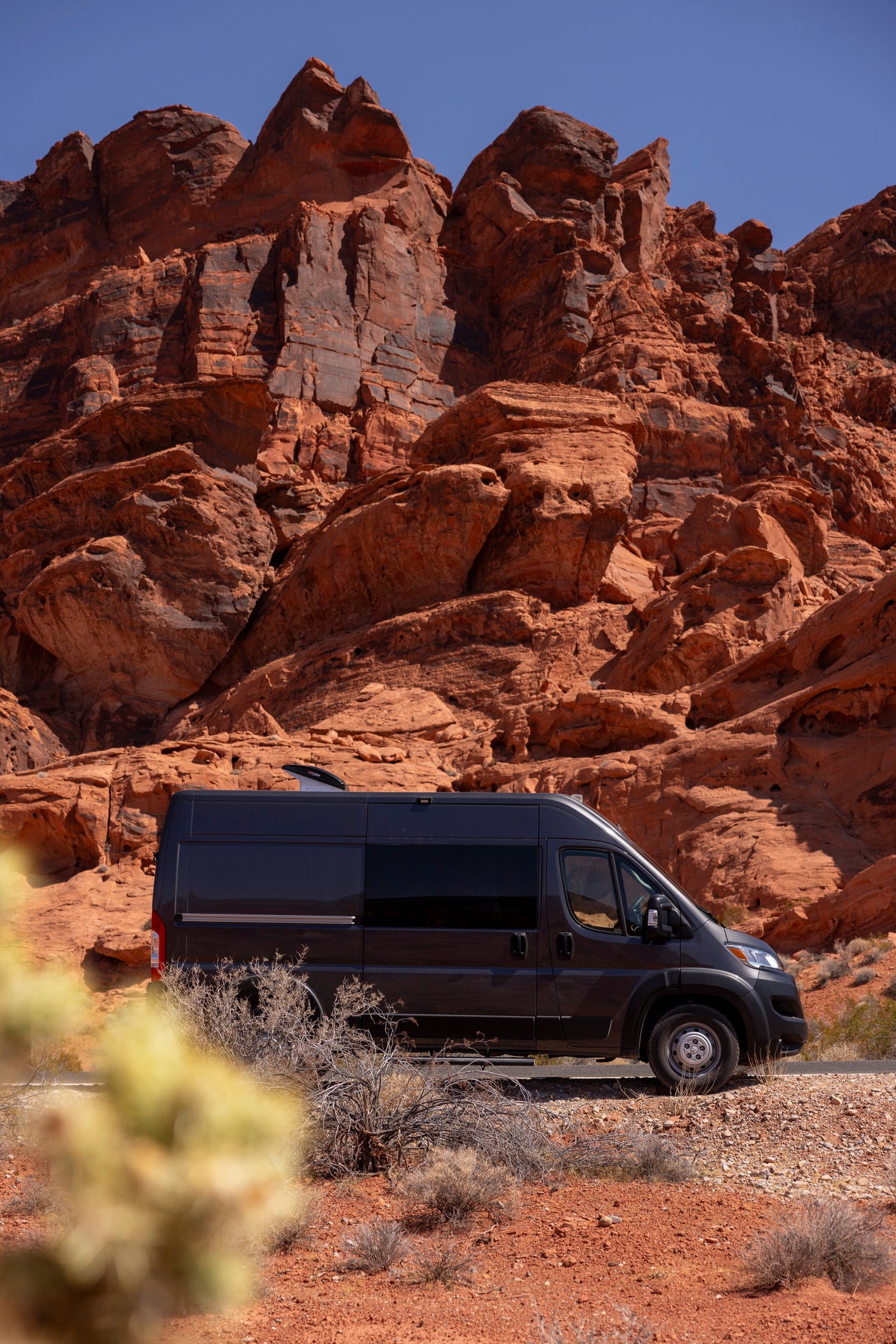 Dark grey RV rental parked among majestic red rock formations in a sunny desert landscape under a clear blue sky.