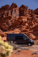 Dark grey RV rental parked among majestic red rock formations in a sunny desert landscape under a clear blue sky.