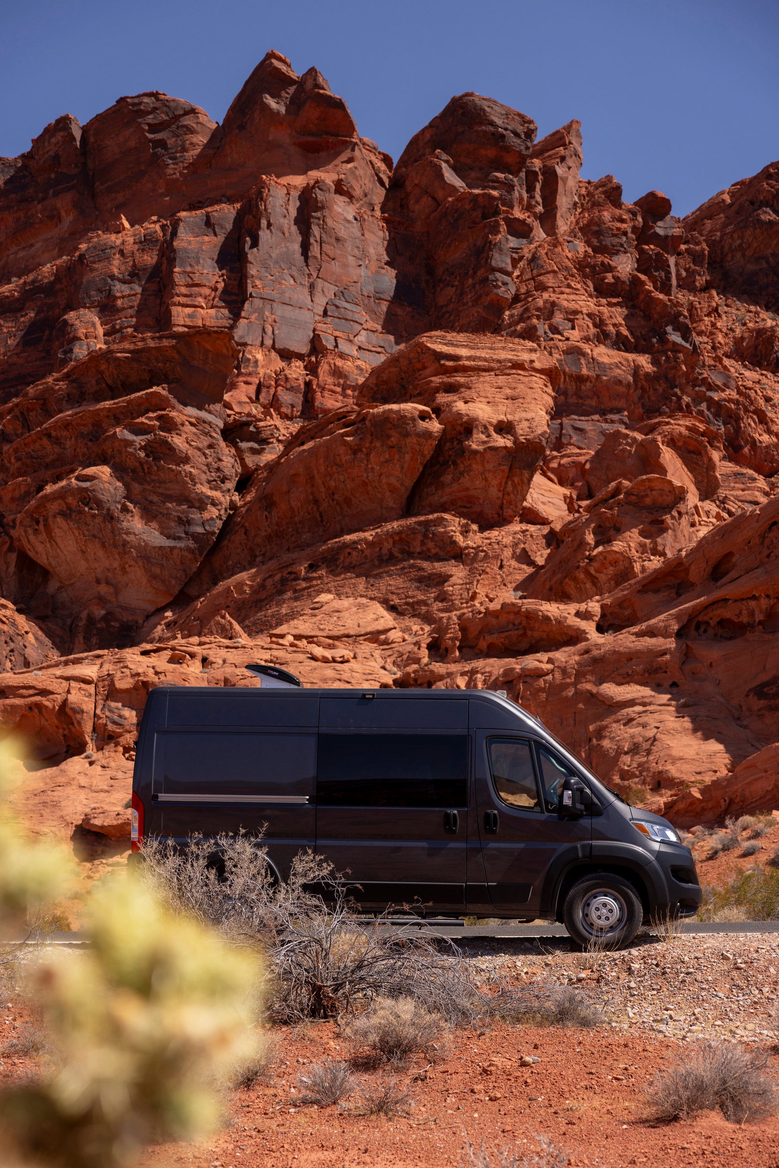 Dark grey RV rental parked among majestic red rock formations in a sunny desert landscape under a clear blue sky.