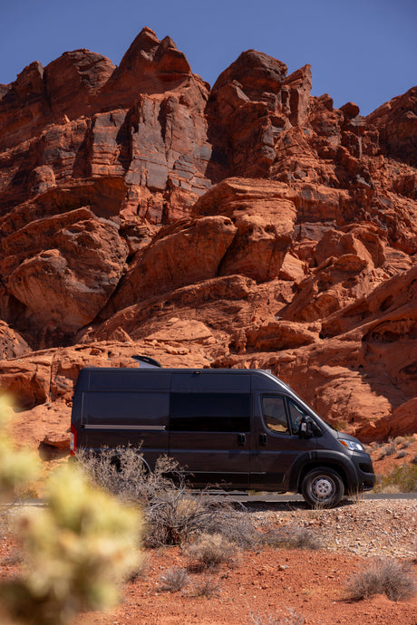 Dark grey RV rental parked among majestic red rock formations in a sunny desert landscape under a clear blue sky.