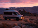 A silver Mercedes Sprinter rv rental, a Tranquility 4x4, rests in a desert landscape with snow-capped mountains at sunset.
