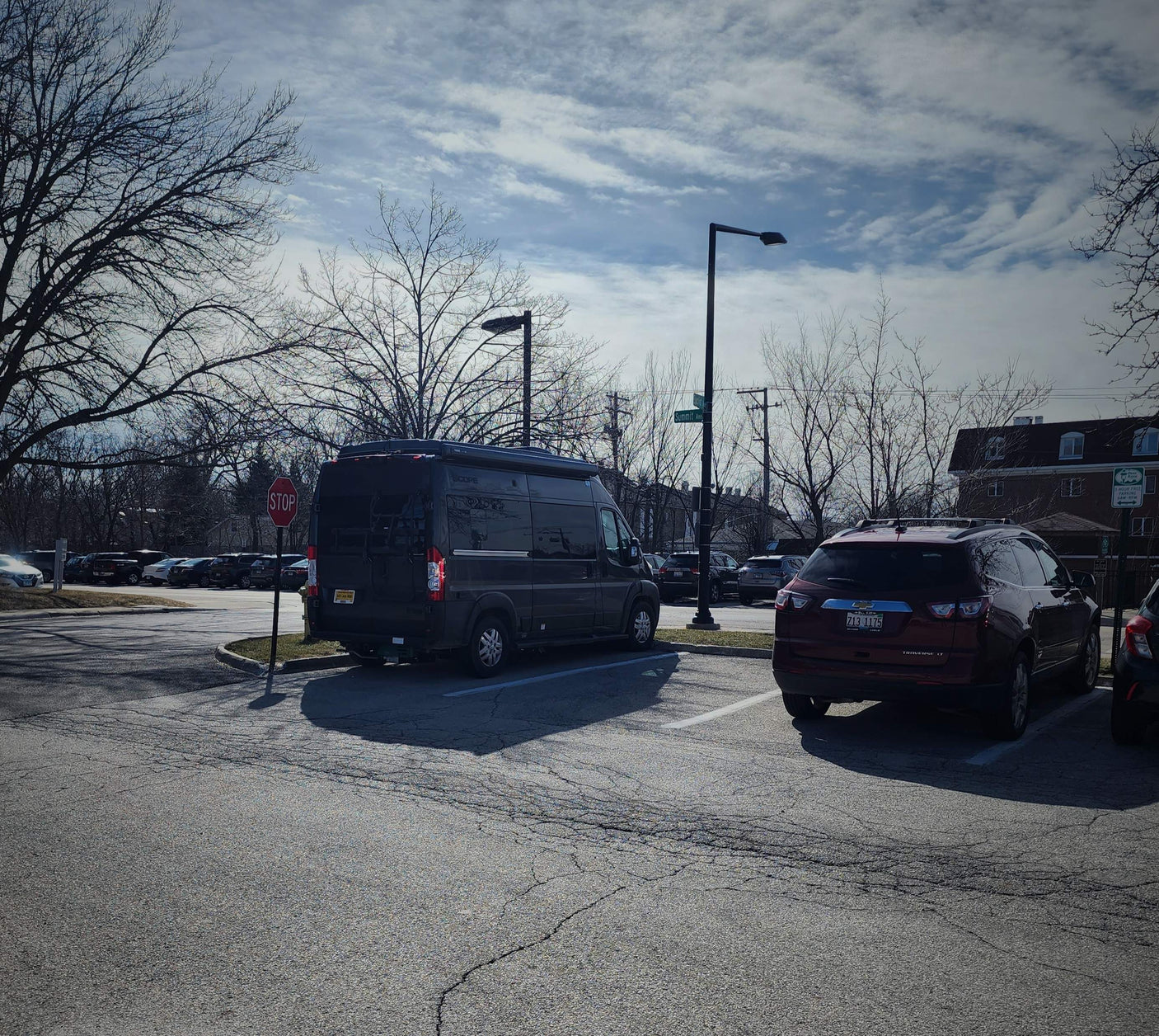Dark gray camper van, an RV rental, parked in a lot with a red SUV and bare trees under a cloudy sky.