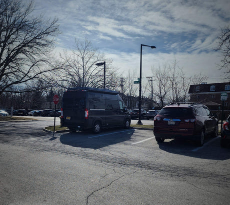 Dark gray camper van, an RV rental, parked in a lot with a red SUV and bare trees under a cloudy sky.
