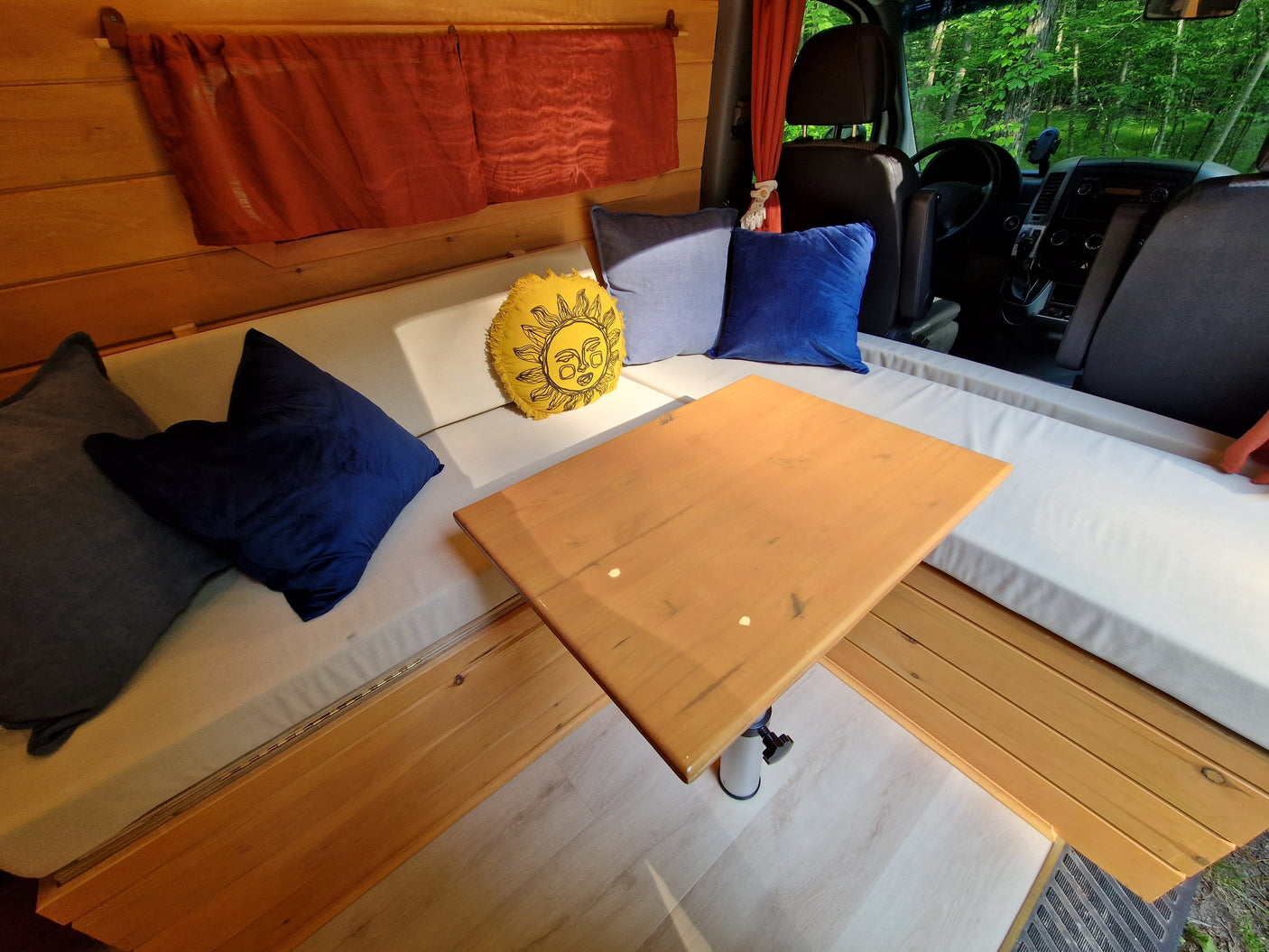 Cozy rv rental interior: a comfy camper setup with wooden walls, padded benches, pillows, a central table, and a lush forest view.