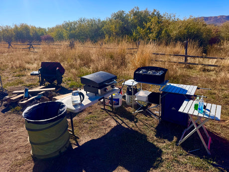 Outdoor camp kitchen setup for an RV rental trip: portable griddle, stove, tables, and supplies in a sunny field.