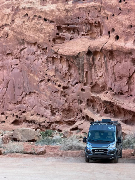 Dark gray RV rental parked on a dirt road in front of an immense, hole-filled red rock wall, showcasing outdoor travel.