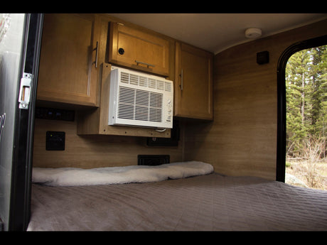 Inside a comfy RV rental: a bed, AC, wooden cabinets, & a window showing a peaceful forest view.