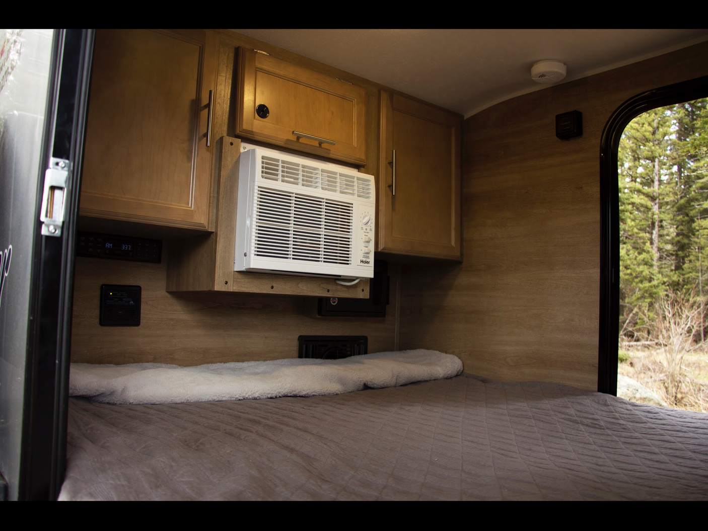 Inside a comfy RV rental: a bed, AC, wooden cabinets, & a window showing a peaceful forest view.
