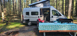 An RV rental camper van with pop-top is set up at a forest campsite with a picnic table and outdoor stove.