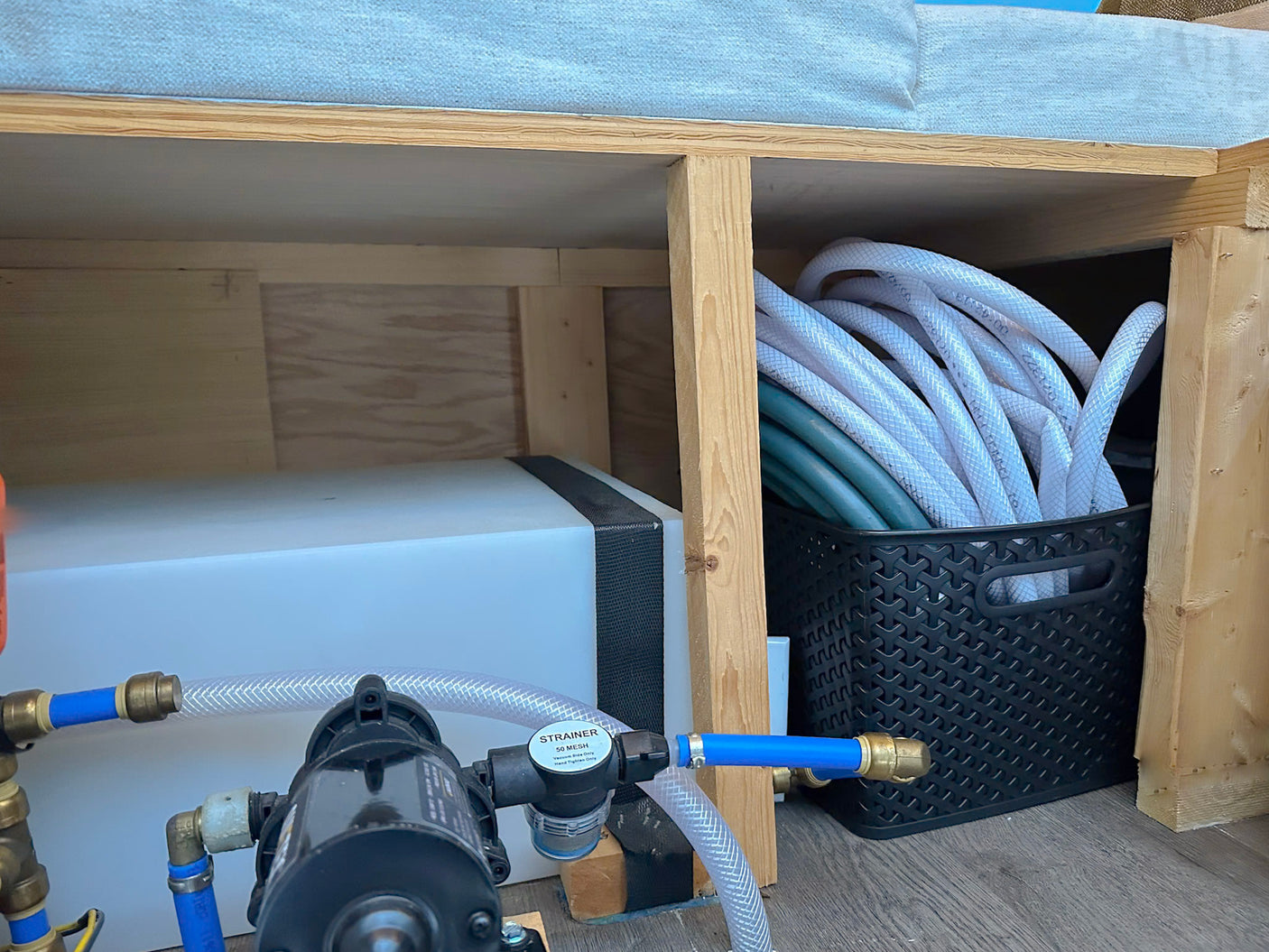 RV rental interior with fresh water tank, pump, and hose connections under a wooden platform, plus a basket of coiled hoses.