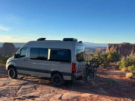 RV rental: Grey Sprinter camper van with bikes on a cliff, overlooking vast red rock canyons & mountains.