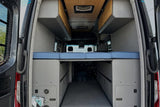 Experience an RV rental: a cozy camper van interior with a raised blue bed, overhead cabinets, and large rear storage.