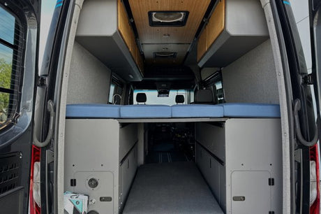 Experience an RV rental: a cozy camper van interior with a raised blue bed, overhead cabinets, and large rear storage.