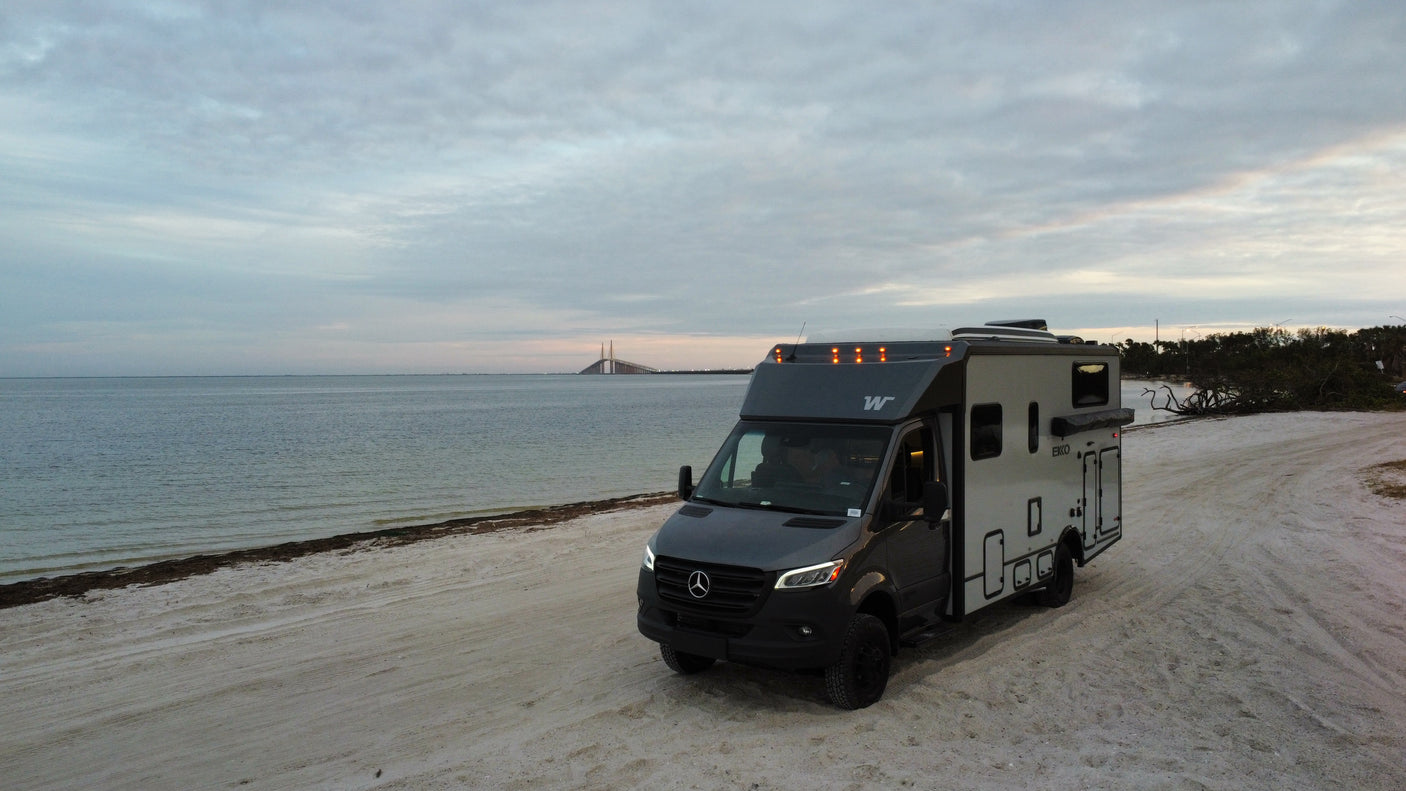 An RV rental, a Winnebago EKO, rests on a sandy beach. A bridge spans the bay under a cloudy sky. Perfect for waterfront travel.